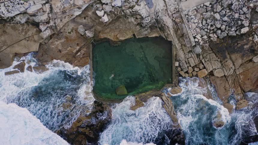 Birds eye view of pool with a person swimming in it on the shore as waves crash in