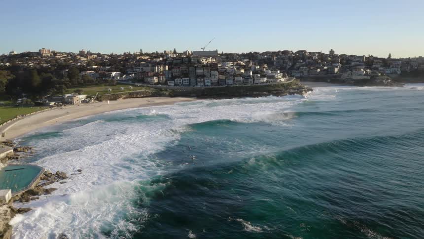 Waves crash on beach seawall and rocks surrounding pool of town under clear blue sky