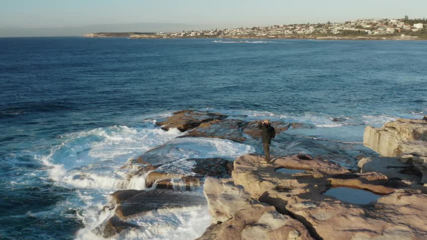 A walks to the edge of a rocky cliff outside of a city and uses a pair of binoculars to look out over the ocean
