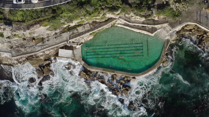 People swim in a large pool on a cliff above the ocean during the day