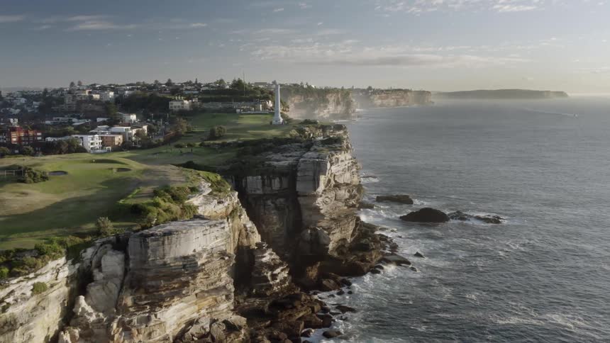 Ocean waves crash into a sheer stone cliff on the edge of a city during the day