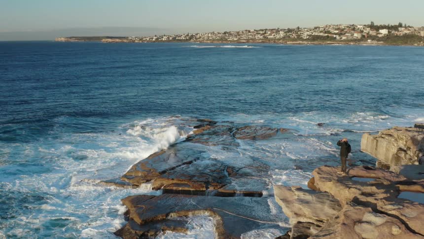 Person walks along rocky outcropping above ocean near coastal town
