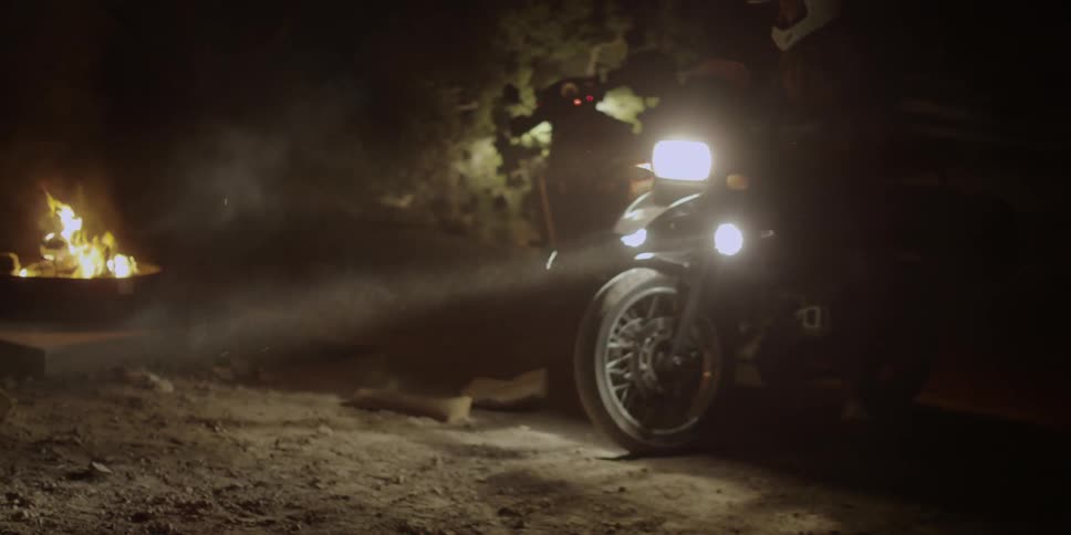 A person on a motorcycle rides up and parks near a campfire