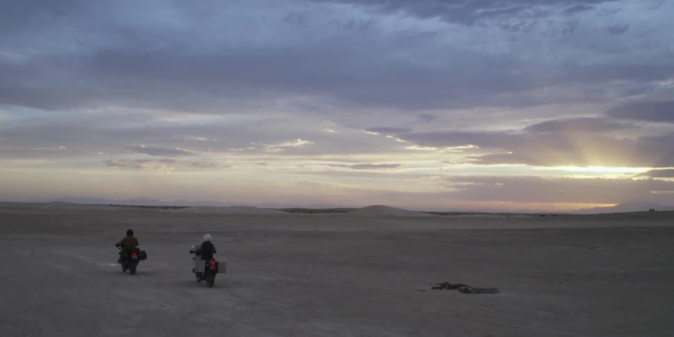 Two people are driving motorcycles over a barren field at sunset