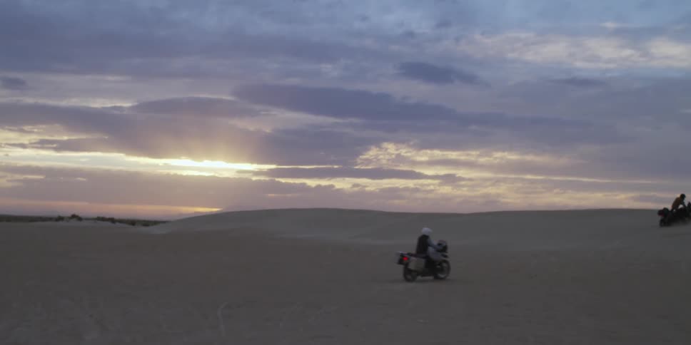 A motorcycle drives over a sand filled area at sunset