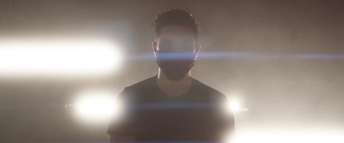 Man wearing black face mask stands back lit by bright lights