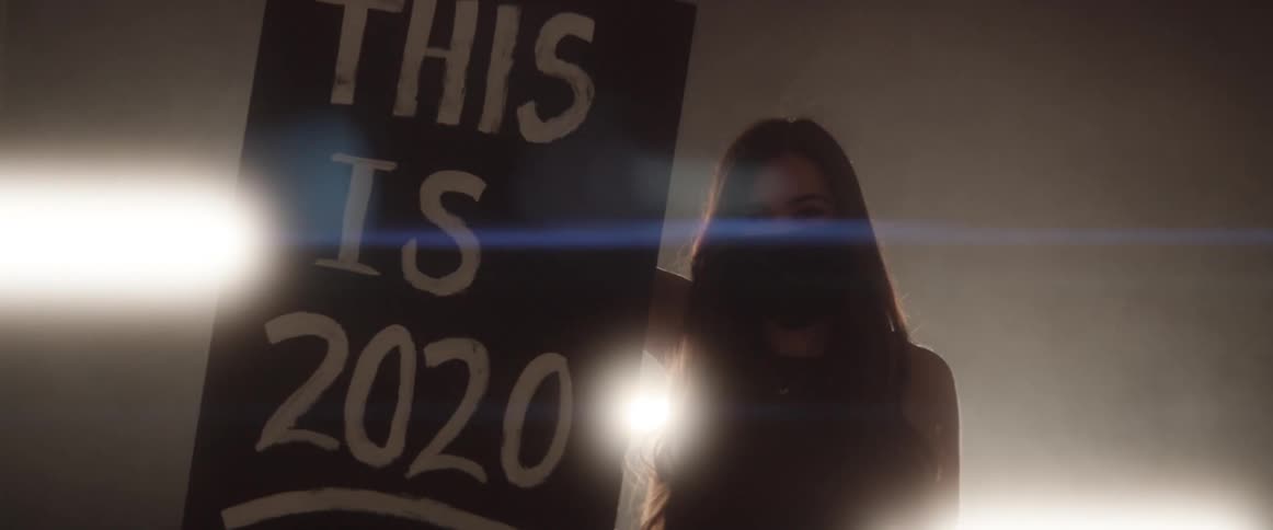 Woman wearing a face mask holds a sign in a dark room in front of strobing fluorescent lights