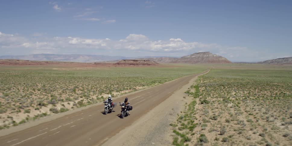Two motorcycles are driving down a small road with a desert on each side