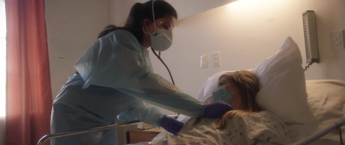 A woman lies in a hospital bed and a nurse listens to her chest