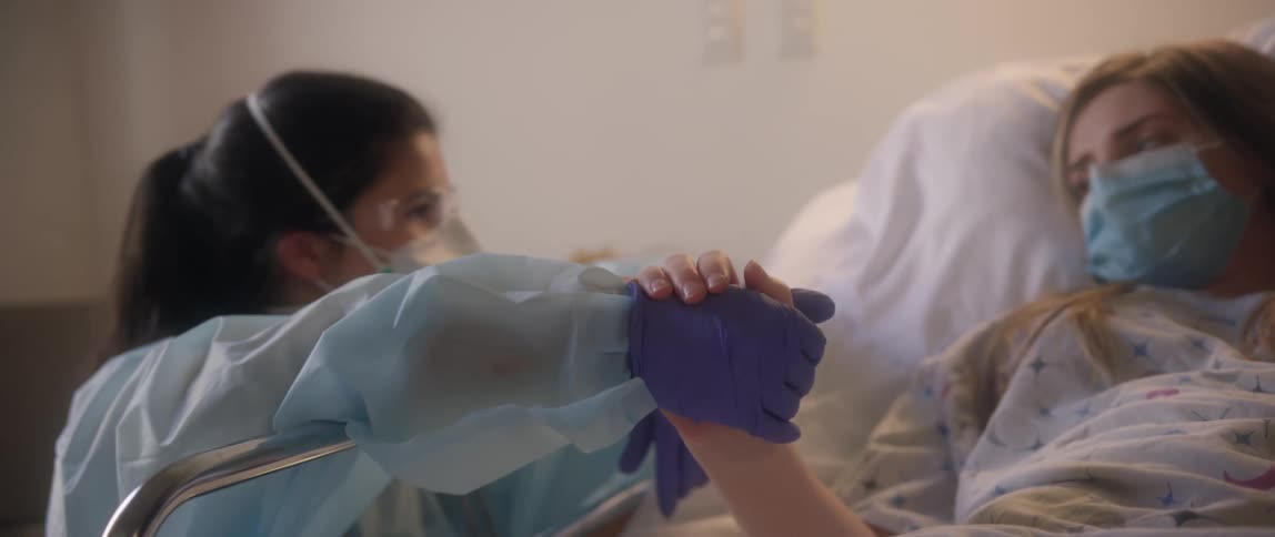 A female nurse kneels beside a female patient and holds her hand