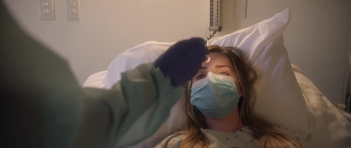 A doctor uses a flashlight to examine the eyes of a coughing patient in a hospital bed