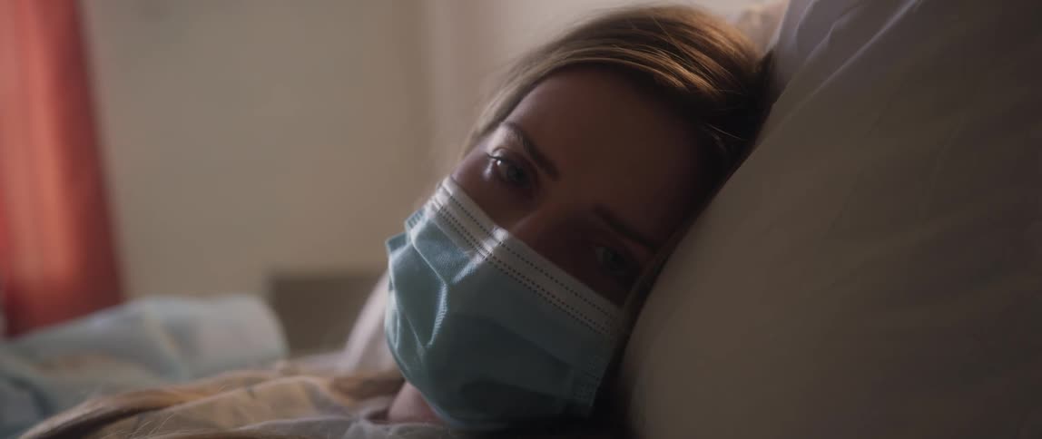A young woman lies in bed at home coughing with a mask on