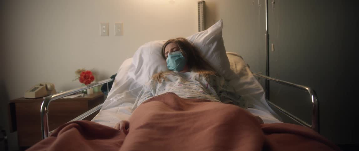A woman in a surgical mask coughs and struggles breathing while laying in a hospital bed