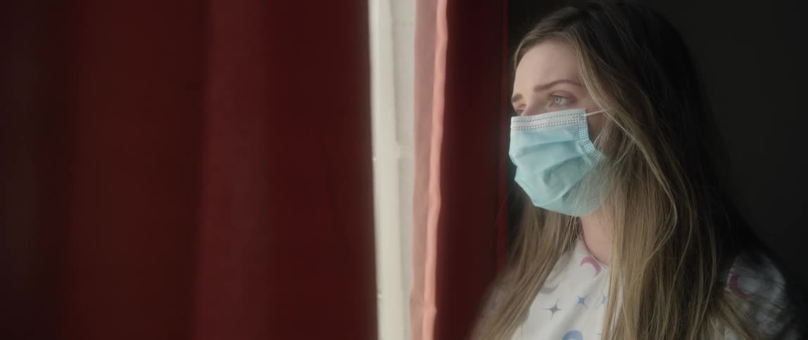 Woman with long brown hair and a blue mask looks out the window at home