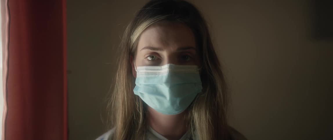A woman in a medical mask looks into the camera while standing near a window in a house