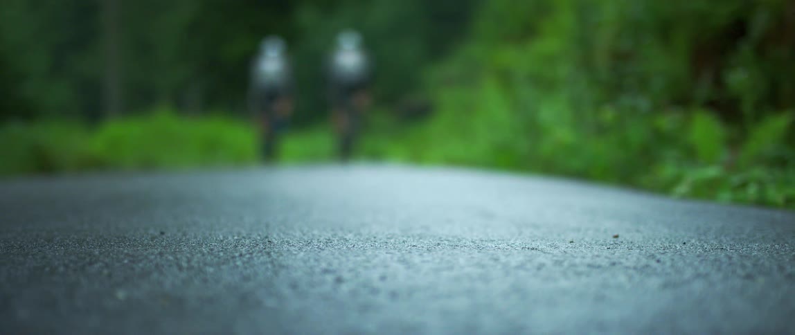 A pair of cyclist ride their bicycles down a wet road through a woodland