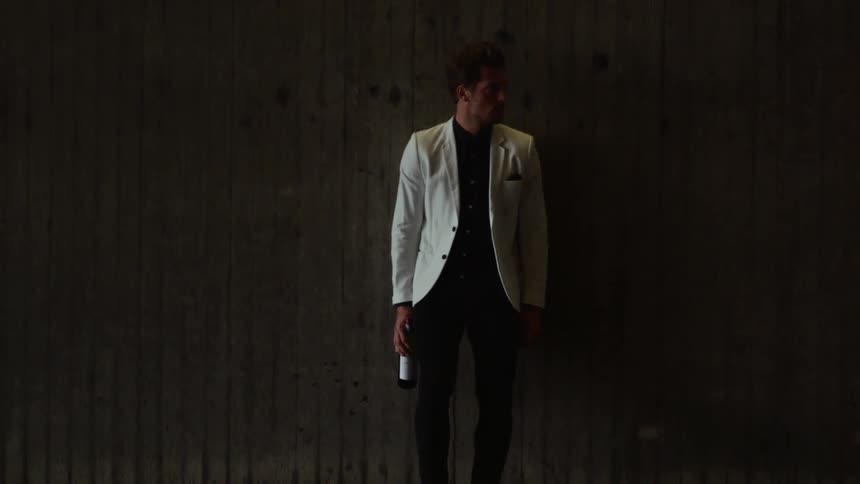 Young man in a white suit holds a wine bottle under a lamp in a dimly lit room