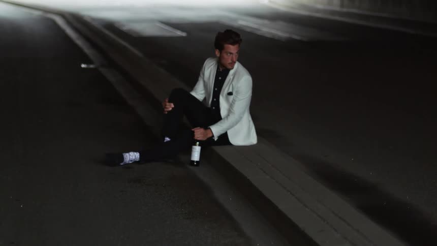 A man in a leisure suit sits on the curb beneath a bridge with a bottle of wine
