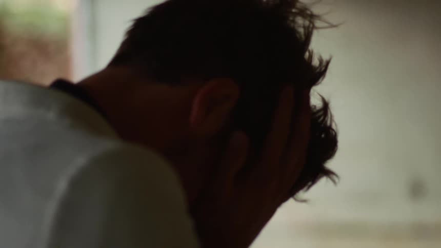 A man at home runs his hands through his hair and sits with his face hidden in his hands