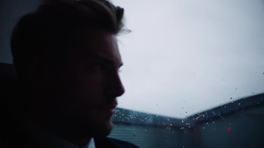 A man with a shirt sitting by a window with the sky and clouds in the background