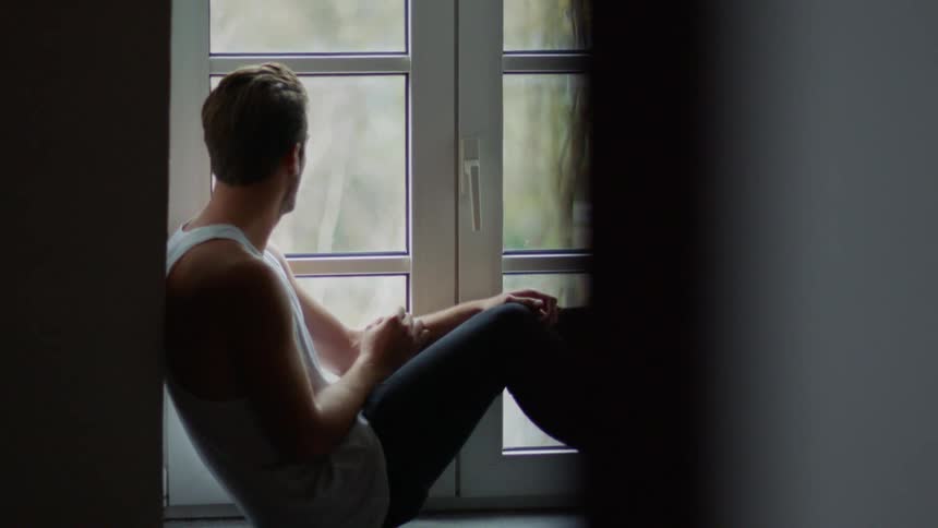 A man sits by a window at home and looks outside