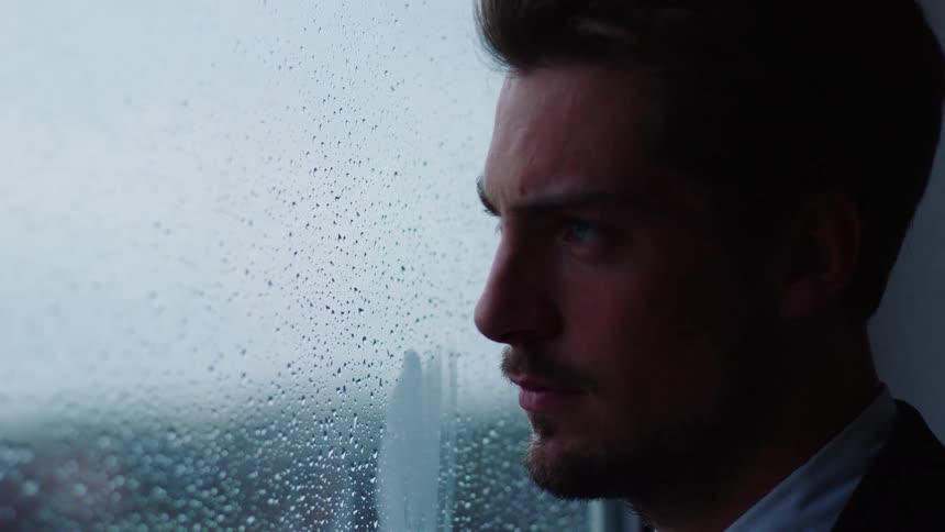 A man in a suit gazes through a window covered in rain drops