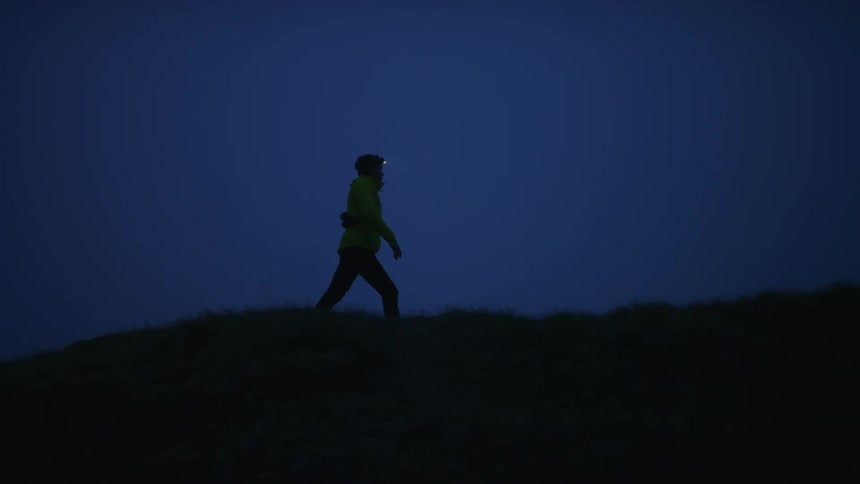 A man with a headlight on walks across the top of a hill at night