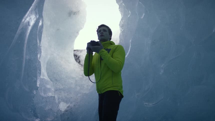 Man standing in an ice cave wearing a bright yellow jacket and dark pants is taking a picture with an opening with the sun shinning through behind him