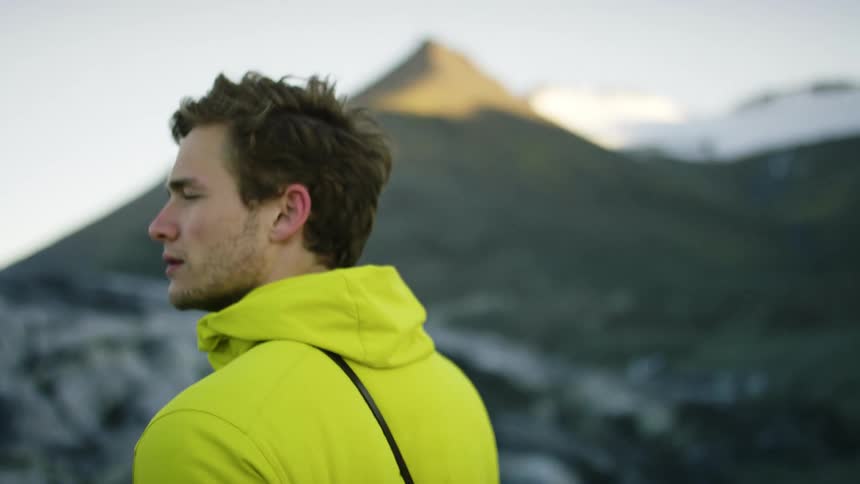 A man with a hoodie looking around with a mountain range the sky and clouds in the background
