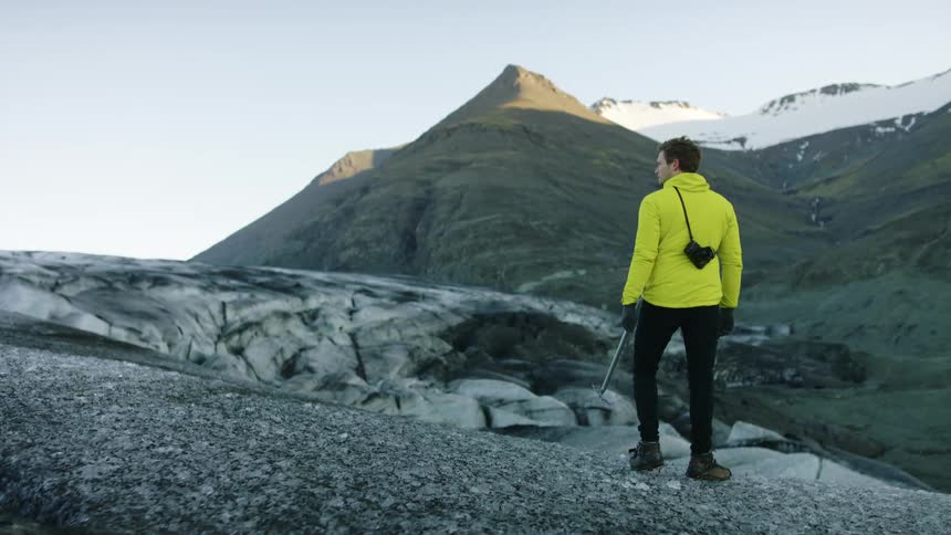 A man with a camera and a hoodie standing with a mountain range and the sky in the background