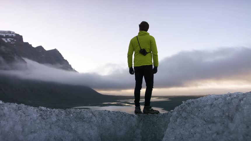 A man stands on top of a glacier and looks out to the mountains and river