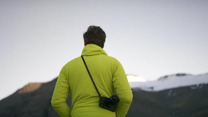 A man with a camera and a hoodie standing with a mountain range and the sky in the background
