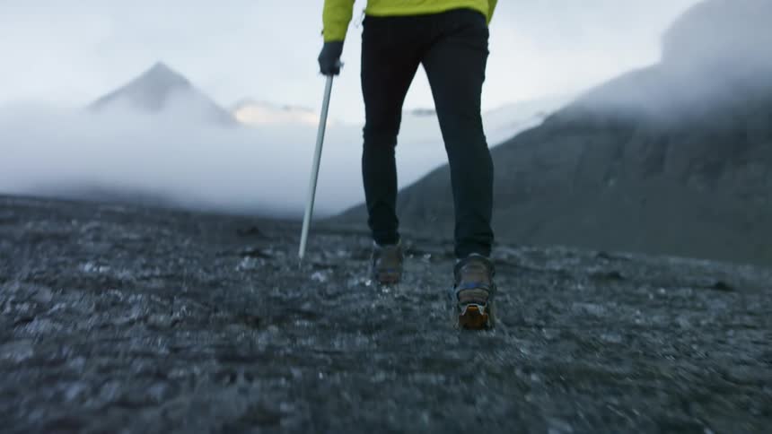 A person in spiked shoes and black pants yellow jacket and black gloves walks on rocky mountain terrain holding a walking stick
