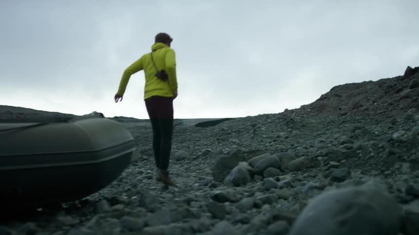 A man carrying a camera around his body jumps out of a small boat that has been parked on a rocky shore