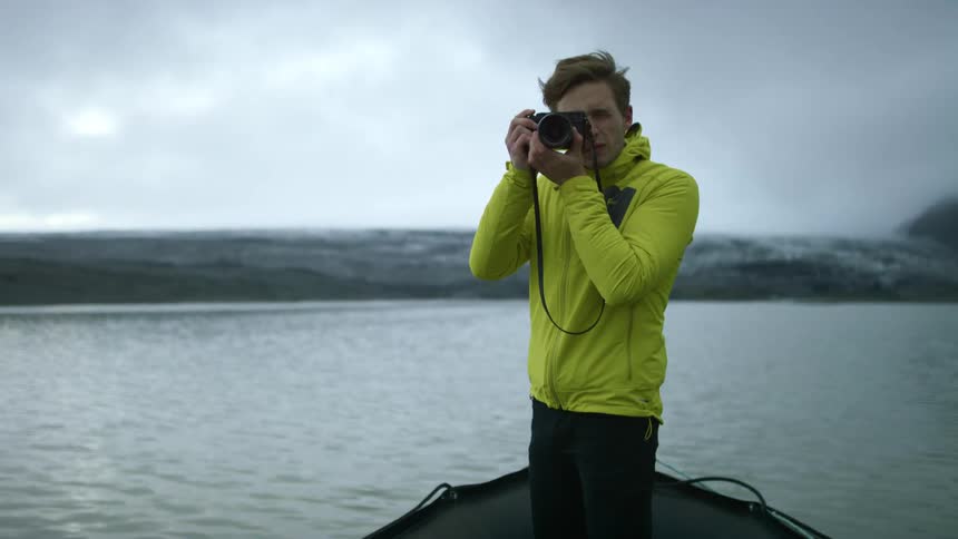 A photographer standing on a small boat as it crosses a lake adjusts his camera before raising it to take a photograph