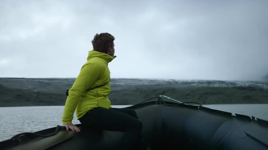 A man sits down on a raft style boat wearing a rain jacket and has a camera strapped around his shoulder