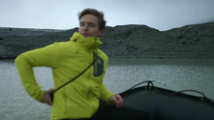 A man wearing a camera around his chest rides a boat towards the gray soil of the shore