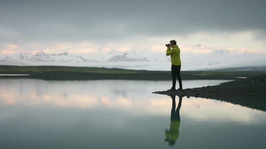 A photographer standing on the shore of a lake raises his camera to take a photographer