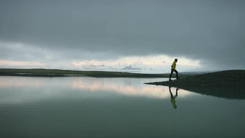 A man looking out over a calm lake from the shore on a cloudy day turns and walks away