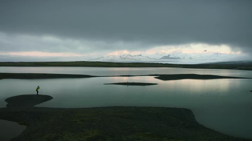 A person crouches next to a lake with snowy mountains in the distance