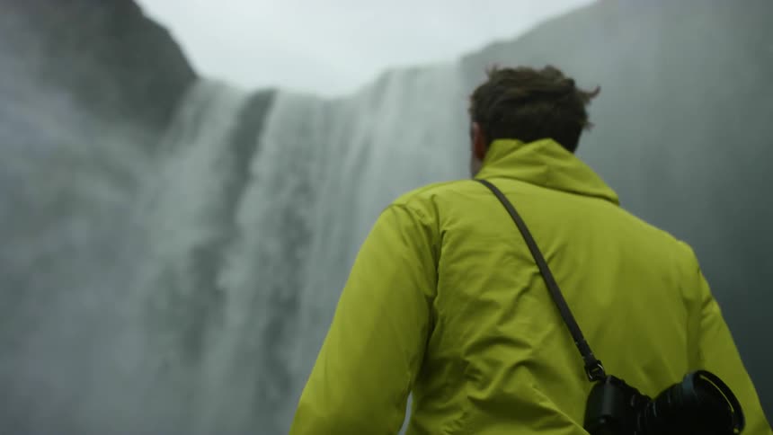 Man wearing a yellow rain jacket walks towards a waterfall while a camera hangs from a strap over his shoulder