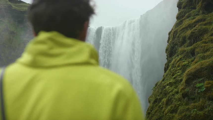 Man wearing a yellow jacket watches a waterfall cascade down the mountain
