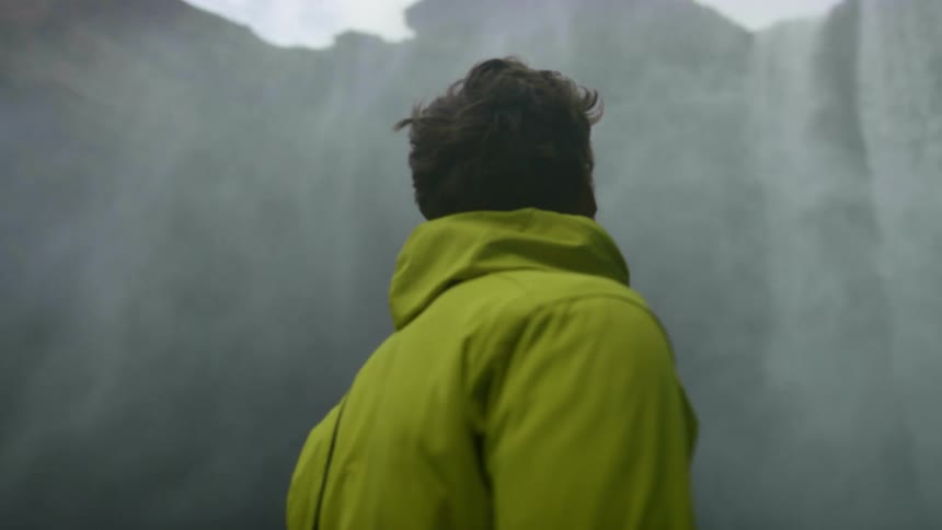 A man standing at the base of a waterfall turns around and looks up at the canyon