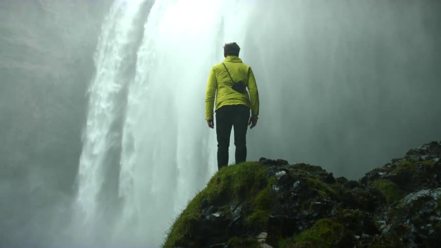 A hiker standing on a mossy rock overlooking a large waterfall in nature