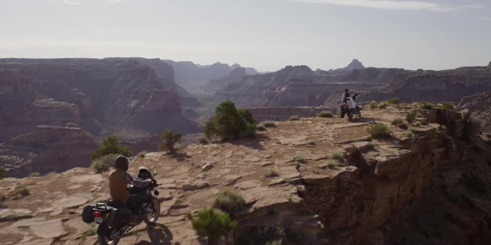 A motorcycle rides up to greet another person on a motorcycle by a cliff