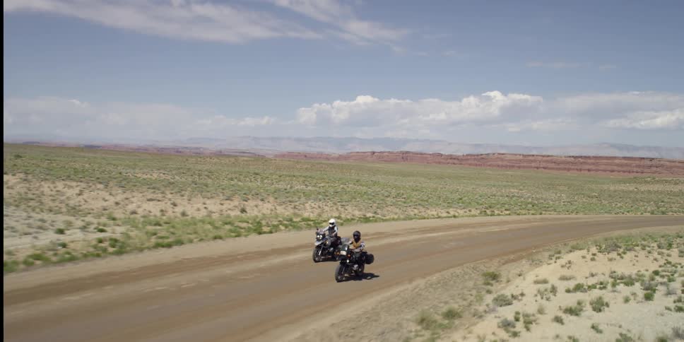 Two motorcycles travel down a dirty road in the desert