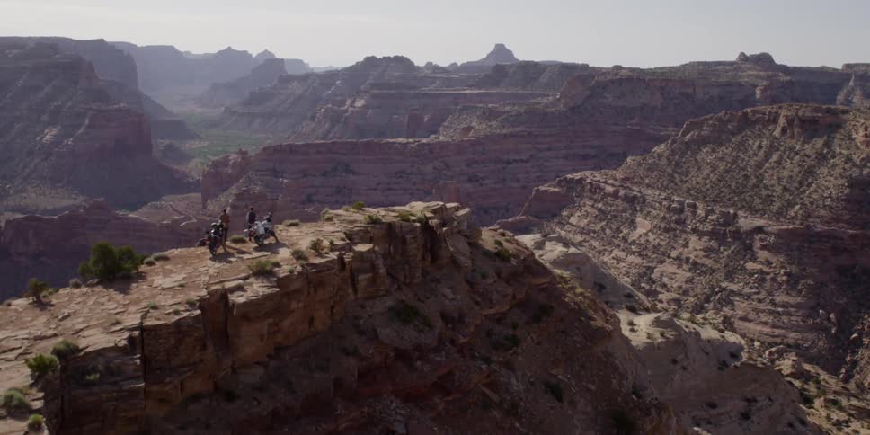 Two men stand near their motorcycles on the top of a cliff in a vast canyon landscape