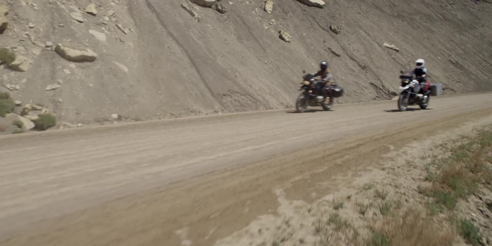 Two motorcycles are driving down a dirt road by a cliff