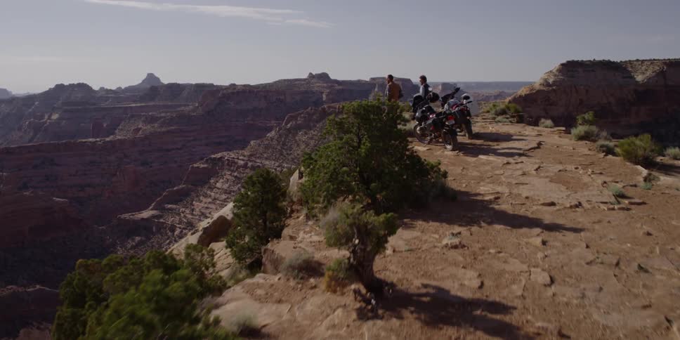 Two men with motorcycles stand on a cliff overlooking a canyon