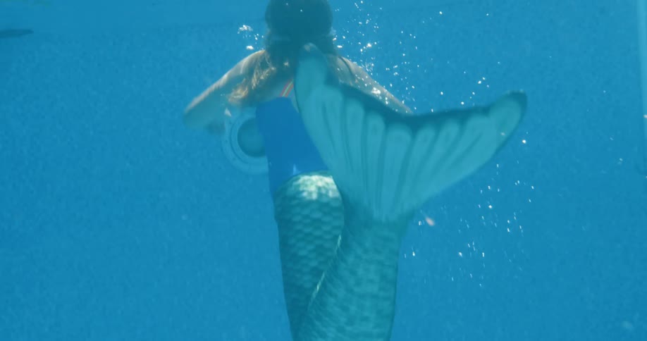 A young girl in a mermaid outfit and googles swims in a swimming pool